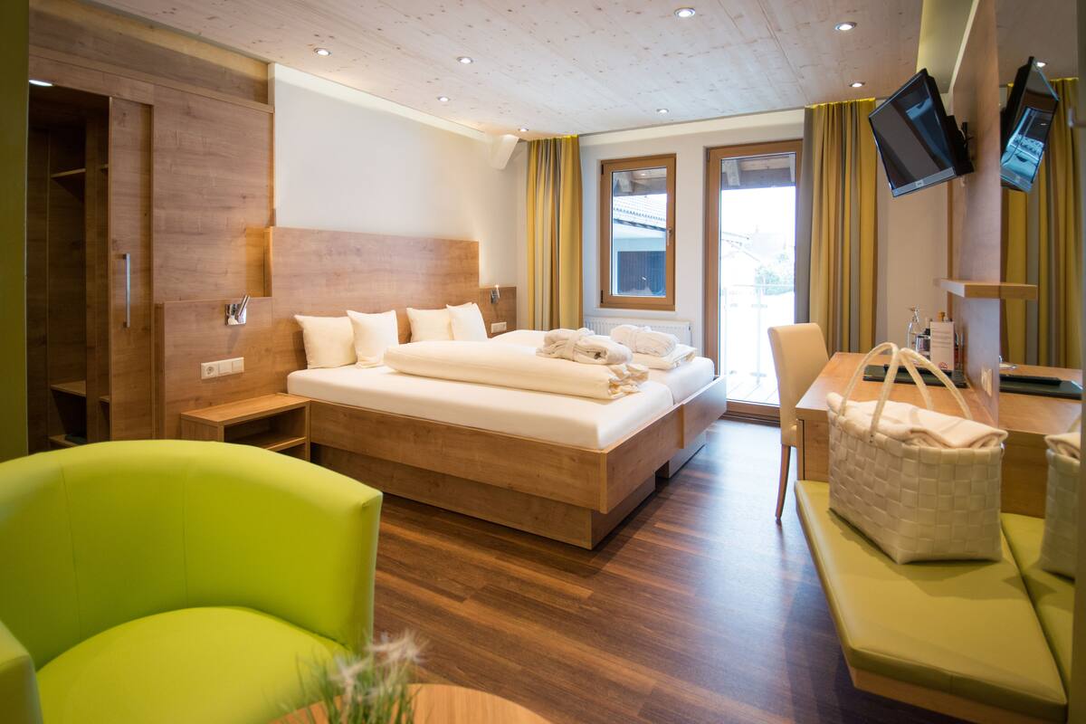 A modern hotel room with a large bed, a desk with a chair, a green armchair, and a TV mounted on the wall.
