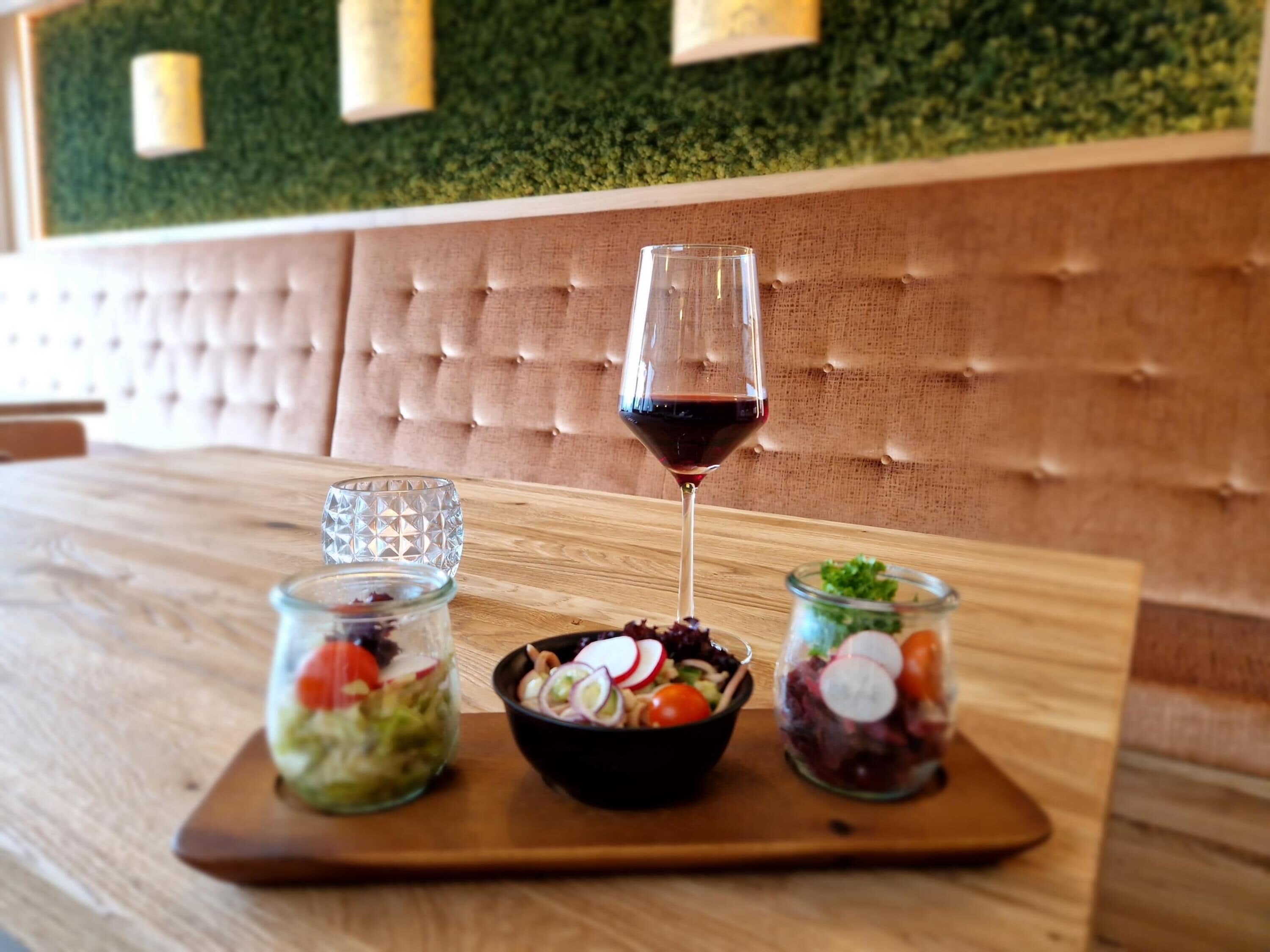 Une table en bois avec un verre de vin rouge, un saladier rempli de salade et deux bocaux en verre contenant les ingrédients de la salade.