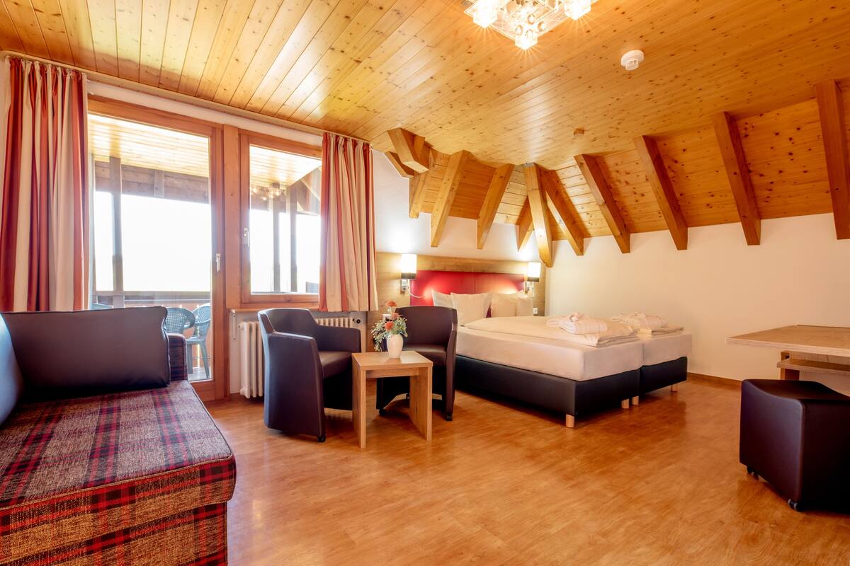 A cozy room with a sofa, a bed, a small table, and a chair. There are windows with curtains, a ceiling with wooden beams, and a radiator.