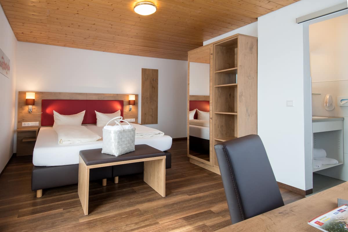 A modern hotel room with a wooden ceiling, a bed with red headboard, bedside lamps, a wooden bench, a desk, and a wardrobe.