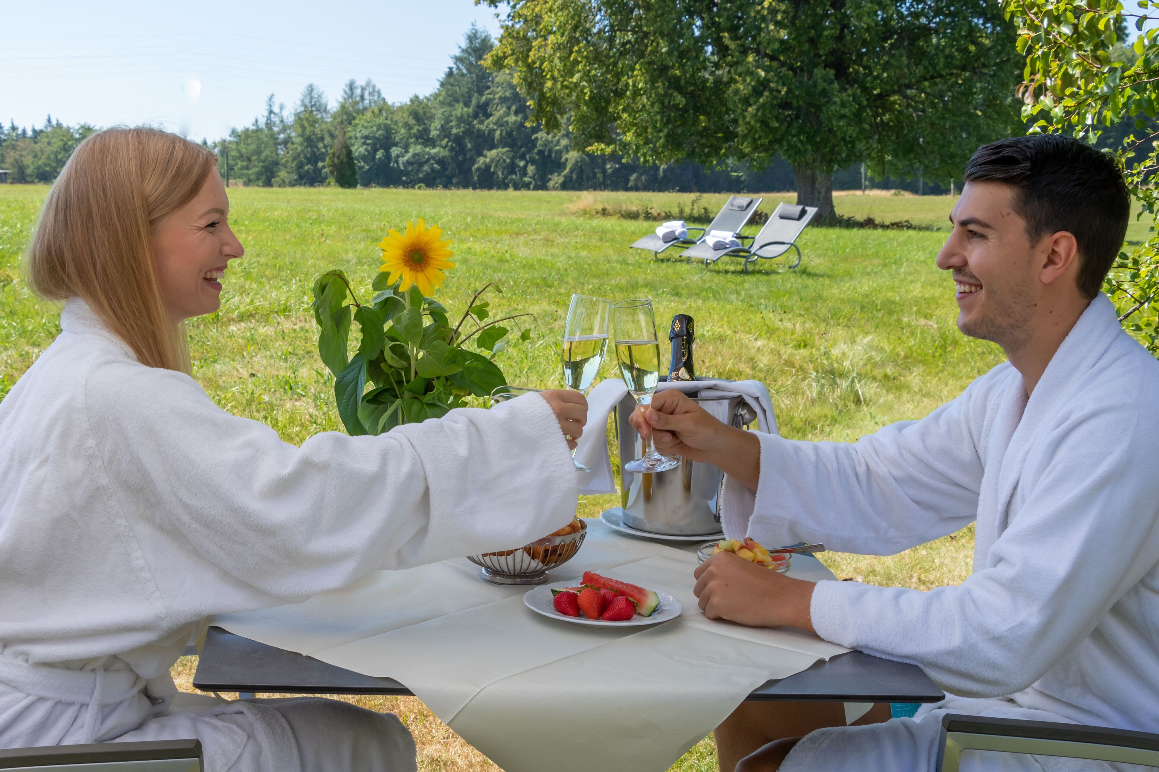 A couple dining outdoors with a table set for two, a bowl of strawberries, a plate of watermelon, and a sunflower.