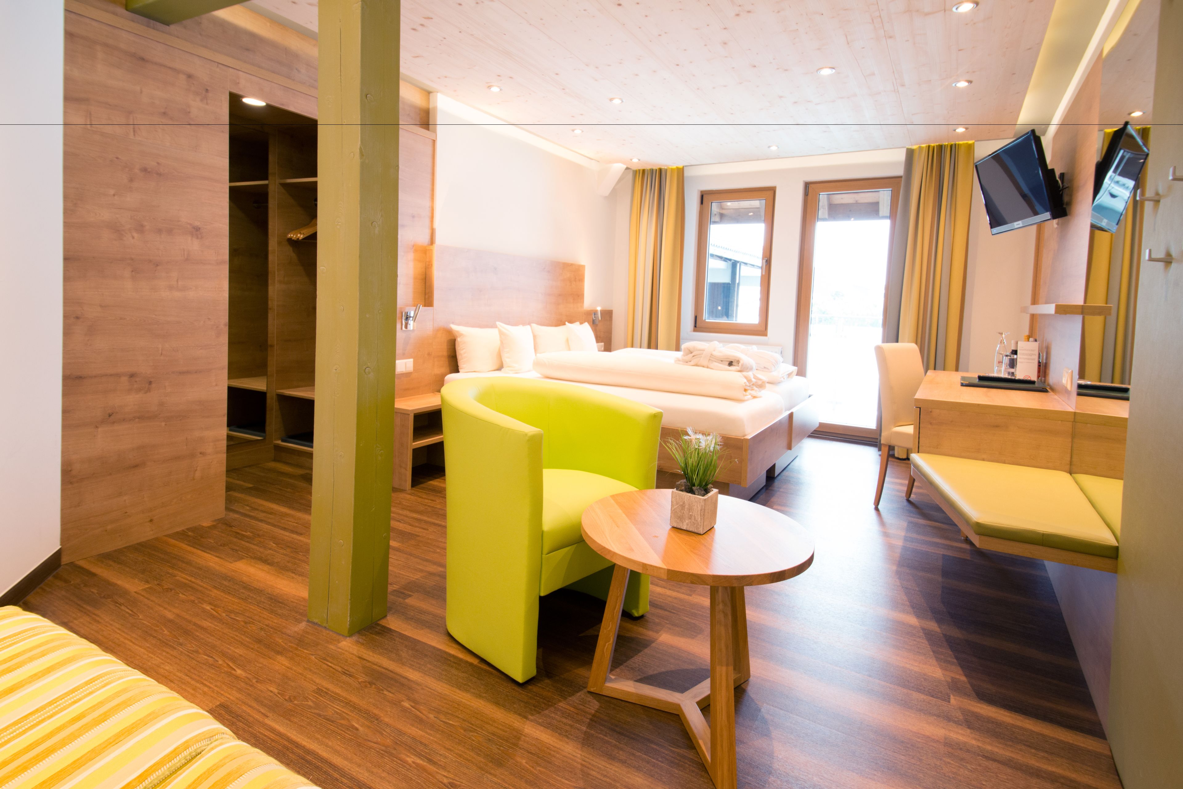 A modern hotel room with a wooden floor, a bed, a green armchair, a small round table, a desk, and a TV mounted on the wall.