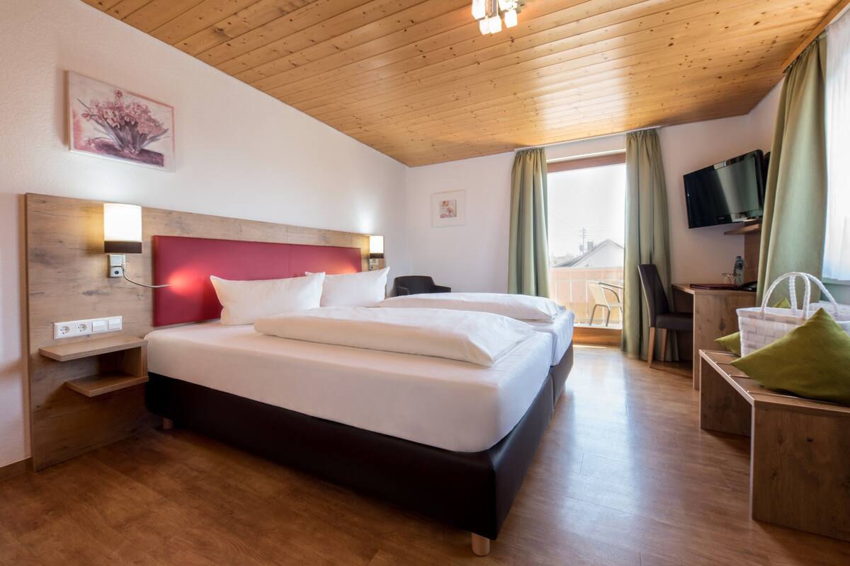 A hotel room with a wooden ceiling, a bed with white bedding, a desk with a chair, and a TV on the wall.
