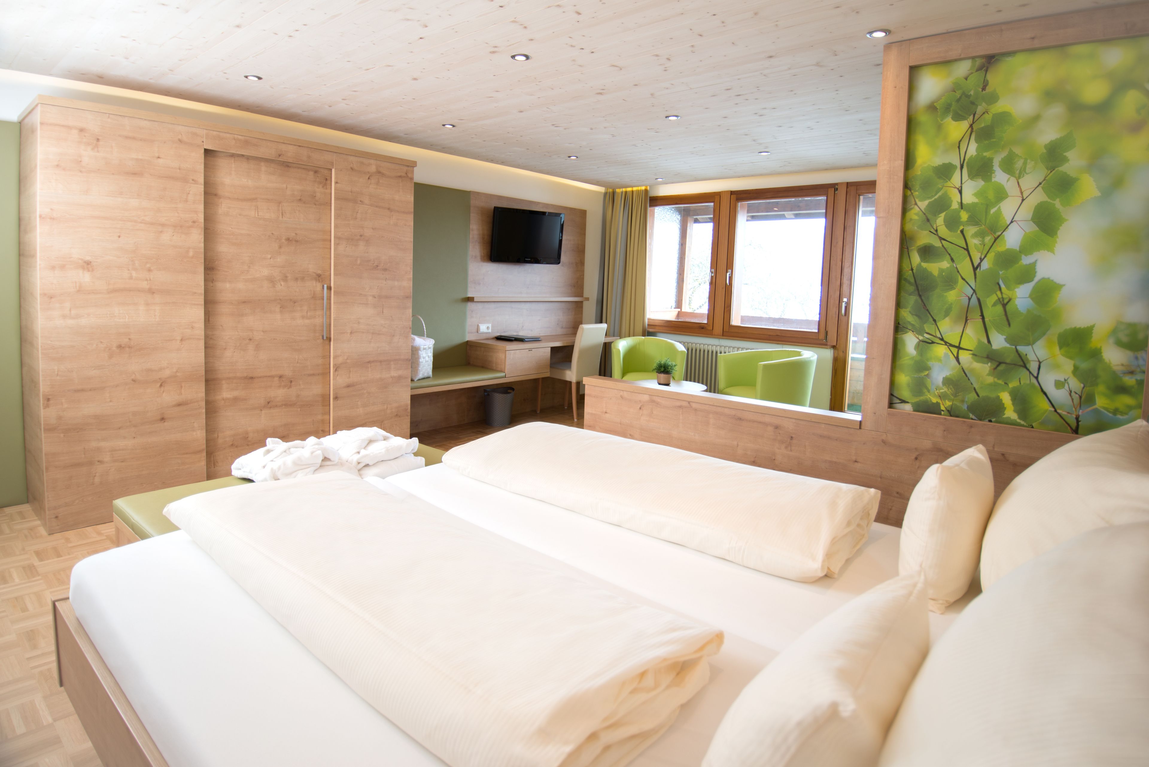 A hotel room with two beds, a wooden wardrobe, a desk with a chair, a television, and a window with a view of greenery.