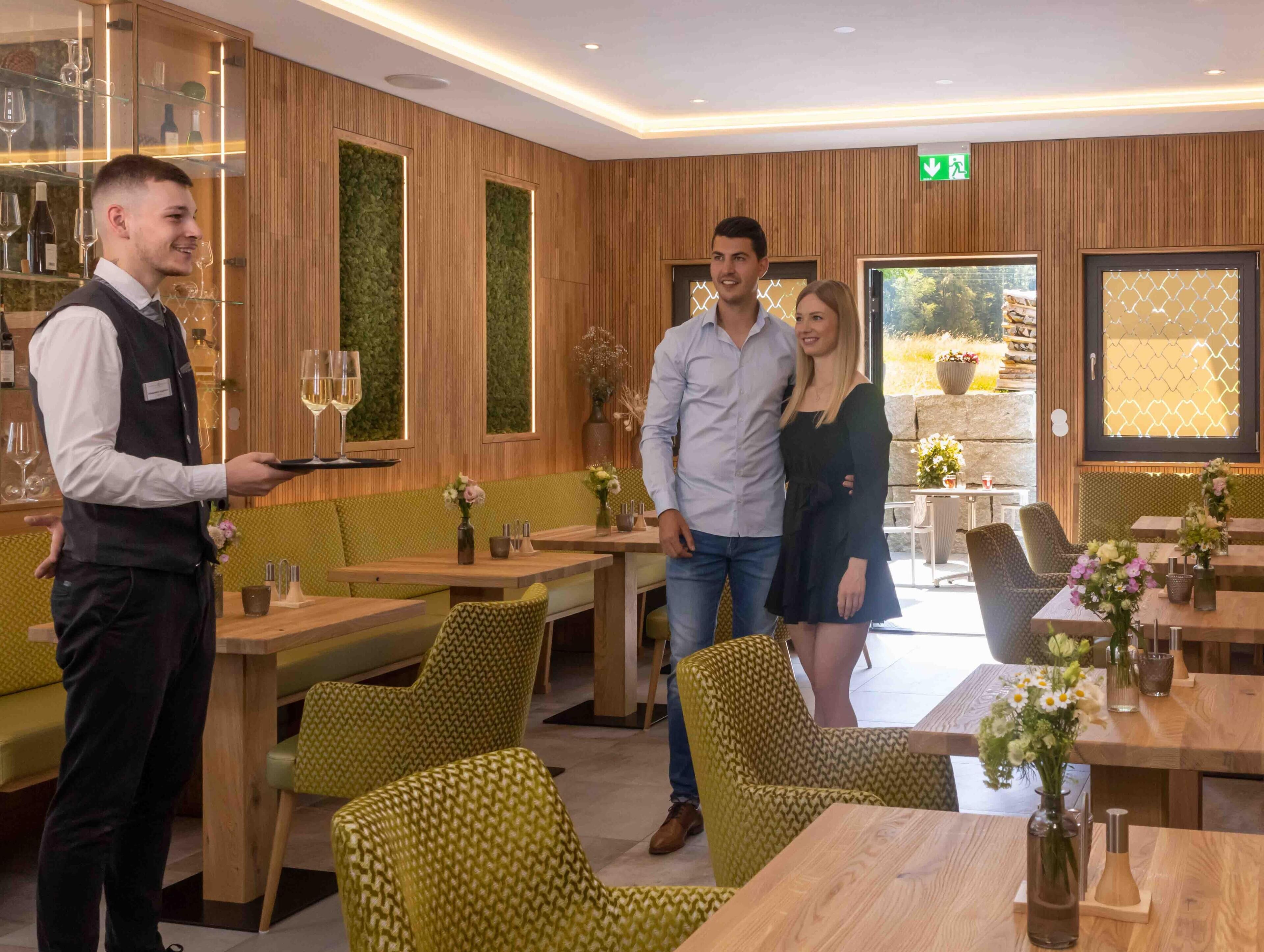 L’intérieur d’un restaurant, avec des lambris en bois, des sièges recouverts d’un tissu vert et un serveur qui apporte un verre de vin.