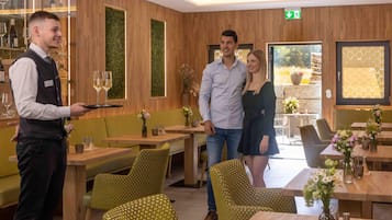 L’intérieur d’un restaurant, avec des lambris en bois, des sièges recouverts d’un tissu vert et un serveur qui apporte un verre de vin.