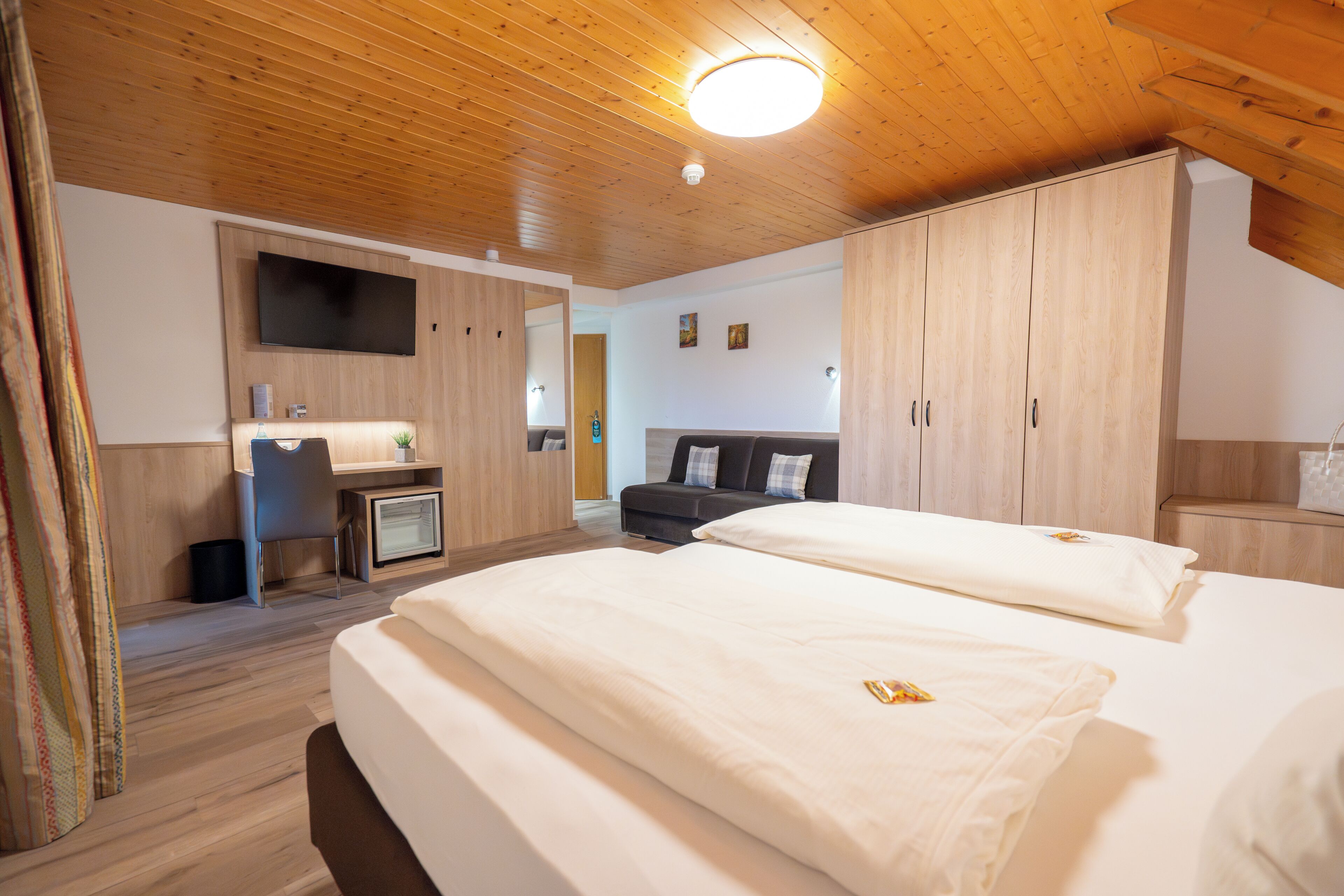 A modern hotel room with wooden ceilings, a flat-screen TV, a desk with a chair, a sofa, and two beds.