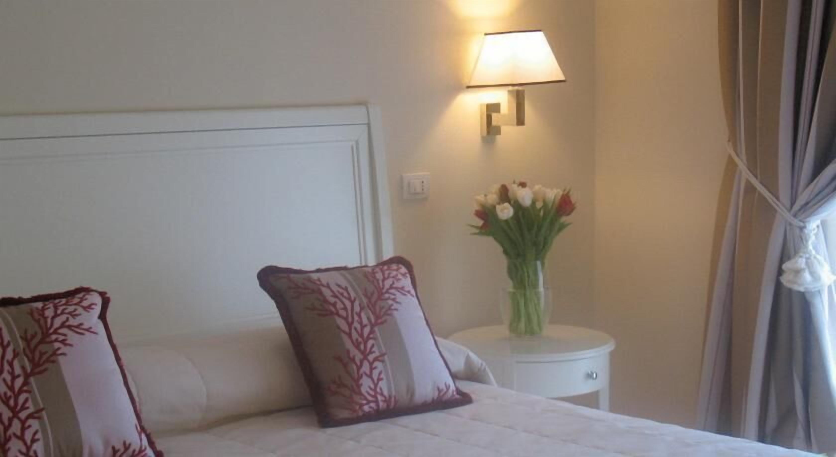 A hotel room with a bed, two decorative pillows, a bedside table with a vase of flowers, and a wall-mounted lamp.