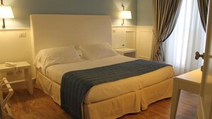 Een net opgemaakt bed met een blauwe deken, witte kussens en een wit hoofdbord, in een kamer met houten vloeren en twee nachtlampjes.