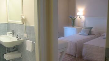 A bathroom with a sink, mirror, and towel rack. There is a bedside table with a lamp and a vase of flowers. Two beds with pillows are visible.