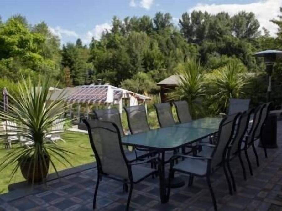 Uma mesa com tampo de vidro e cadeiras, cercada por um jardim com árvores e um gazebo ao fundo.
