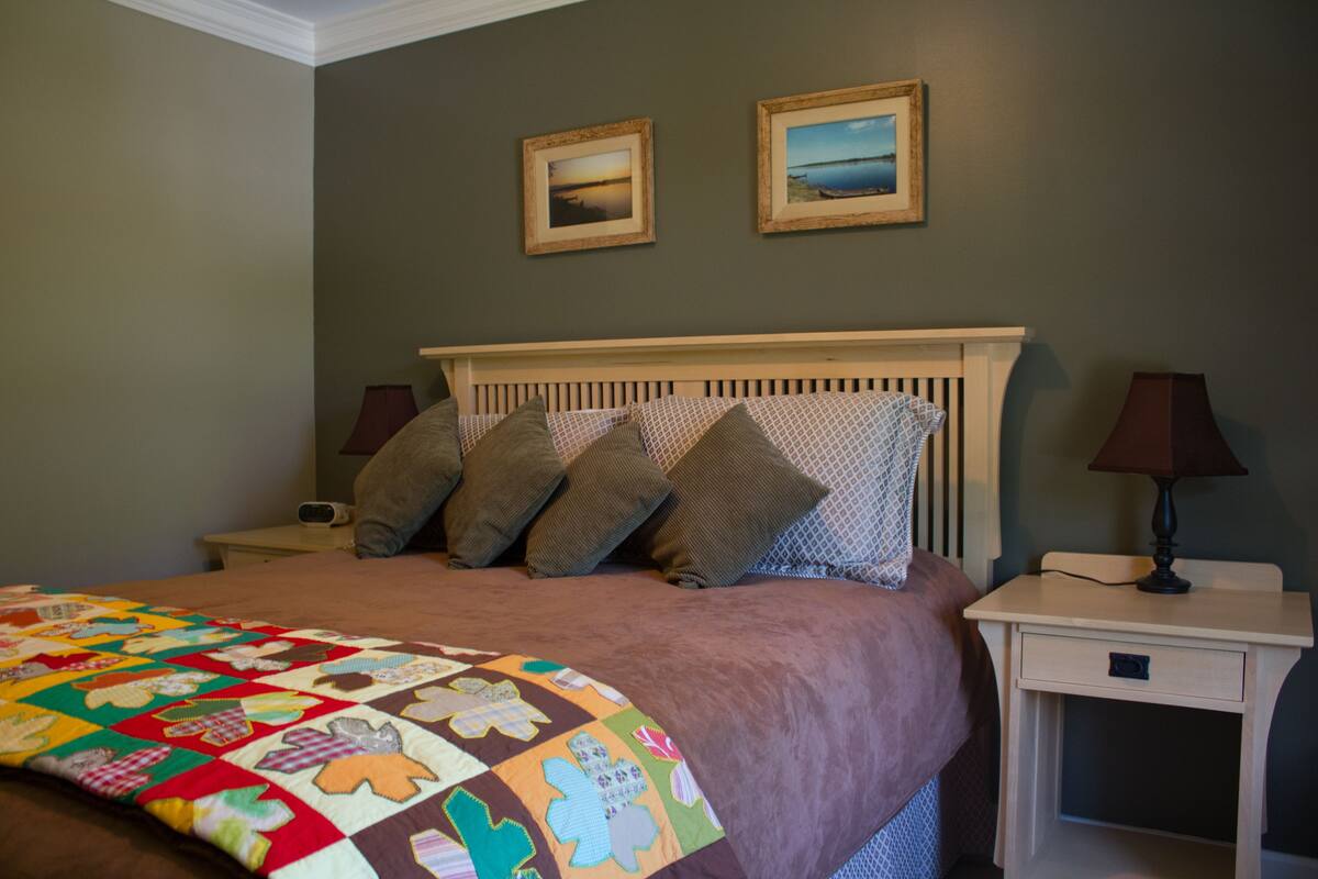 A bedroom with a bed, two bedside tables, a nightstand, a quilt, and two framed pictures on the wall.