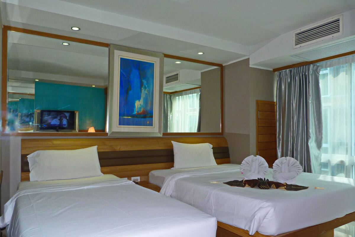 A hotel room with two beds, a television, artwork on the wall, and a large window with curtains.