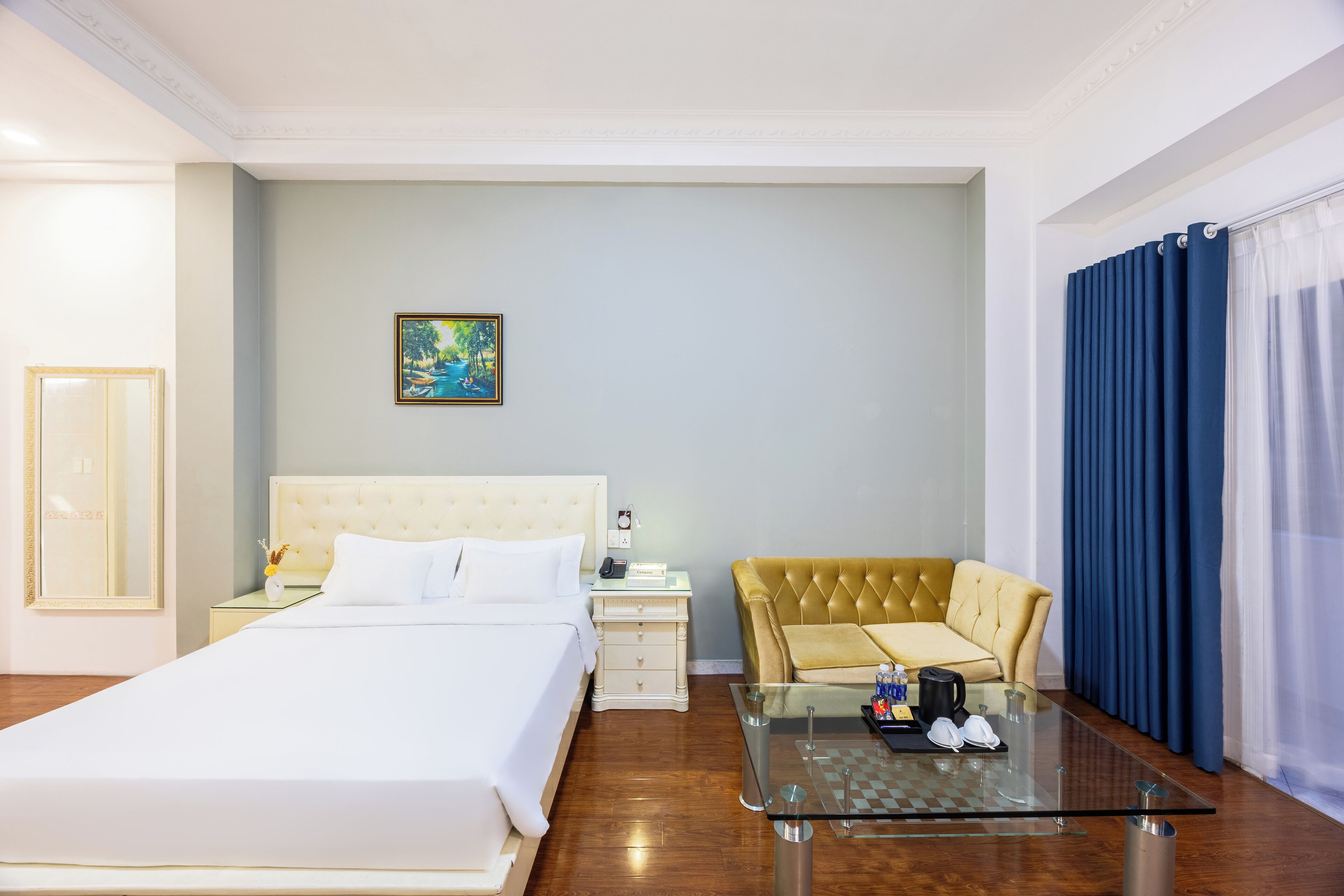A hotel room with a large bed, a glass coffee table, a sofa, a painting on the wall, and a view of the outdoors.