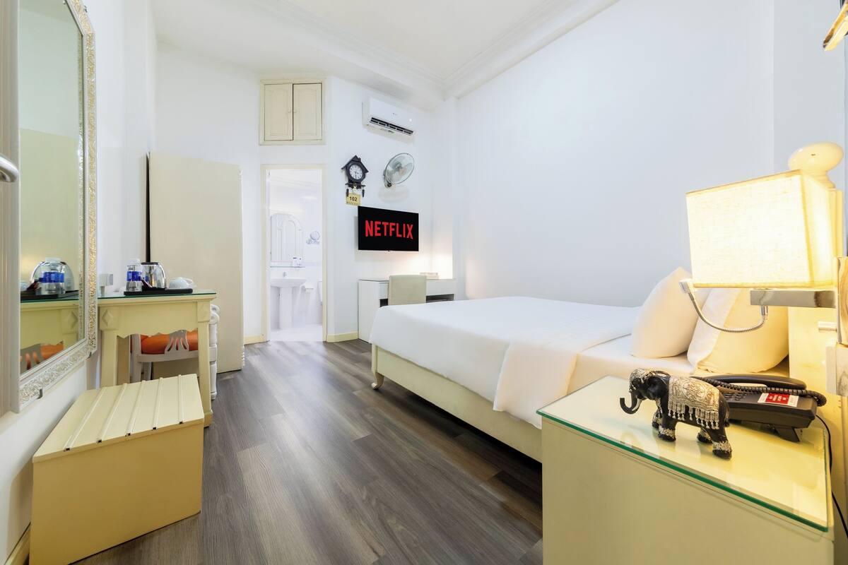 A hotel room with a bed, a bedside table with a lamp, a small elephant figurine, a TV displaying 'NETFLIX', and a mirror.