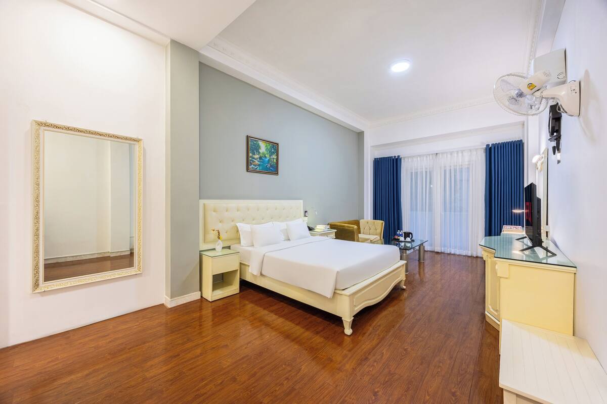 A hotel room with a large bed, a bedside table, a chair, a desk, and a mirror.