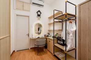 Habitación de hotel compacta con suelo de madera, un escritorio con espejo, una silla, estante con bata y un reloj en la pared.