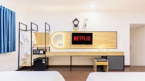 A hotel room with a wooden desk, a television displaying "NETFLIX", a clock, and a fan.