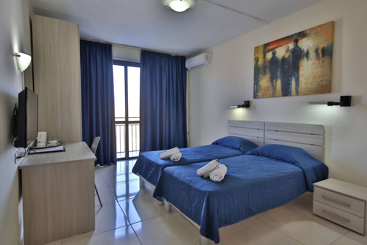 A hotel room with two beds, a desk with a telephone, and a painting on the wall.