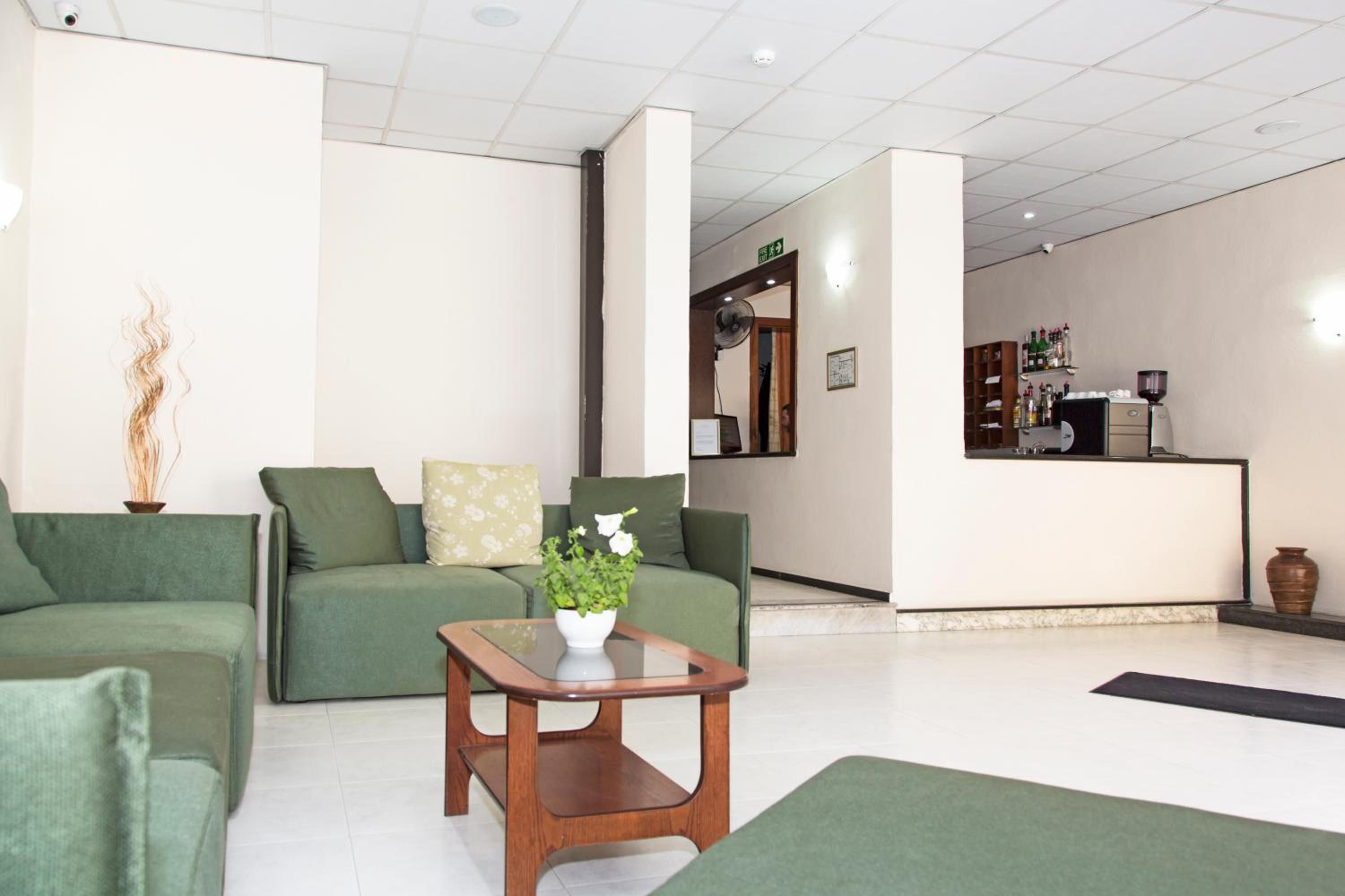 Uma área de recepção com um sofá verde, uma pequena mesa de madeira com uma planta e um balcão de atendimento com diversos objetos.