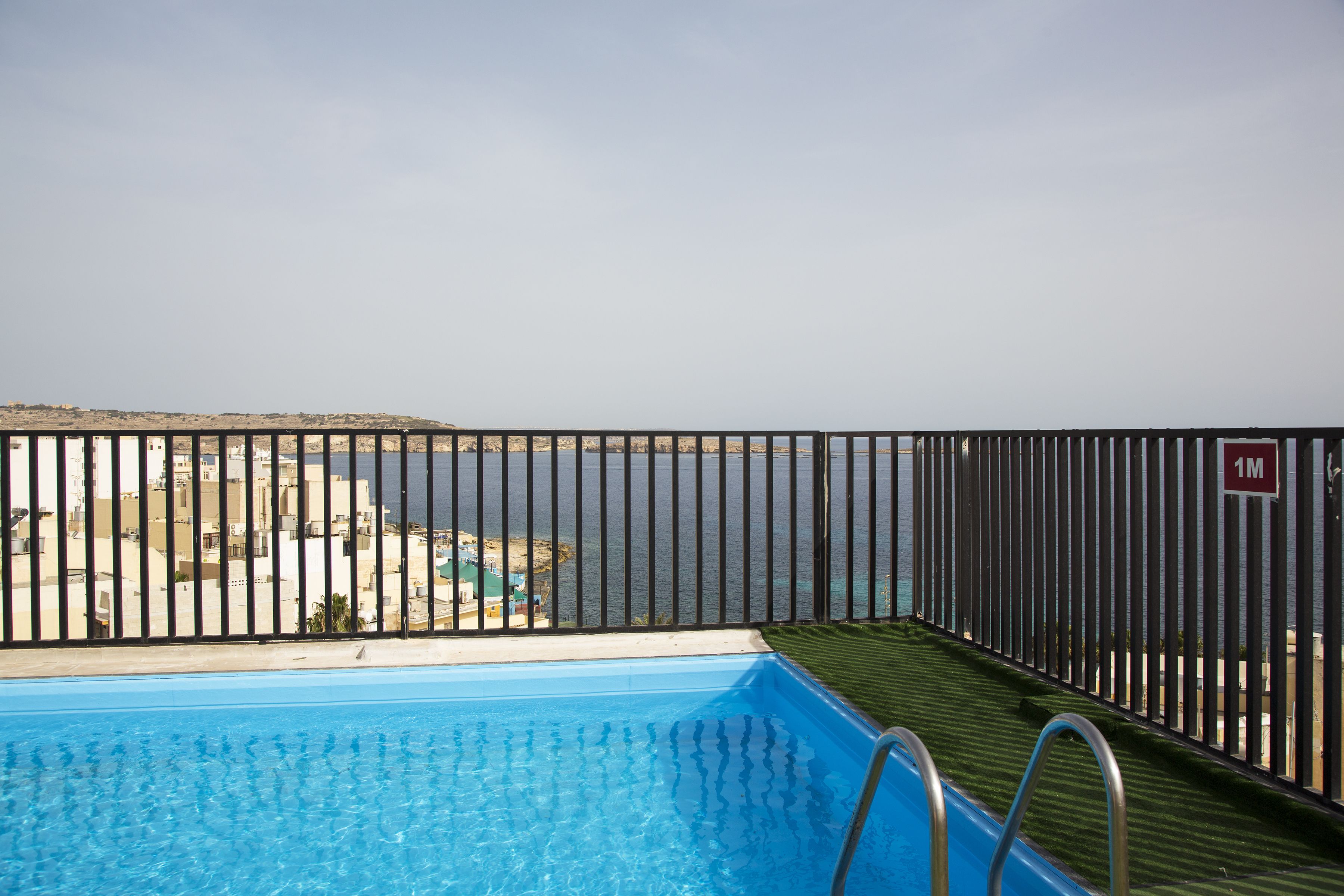 Une piscine bordée d’une rambarde en métal, avec vue sur la mer et, au loin, une côte parsemée de bâtiments.