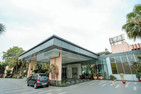 A modern hotel entrance with a covered walkway, palm trees, and a parked car. - Oodles Hotel Chhattarpur South Delhi (New Delhi)