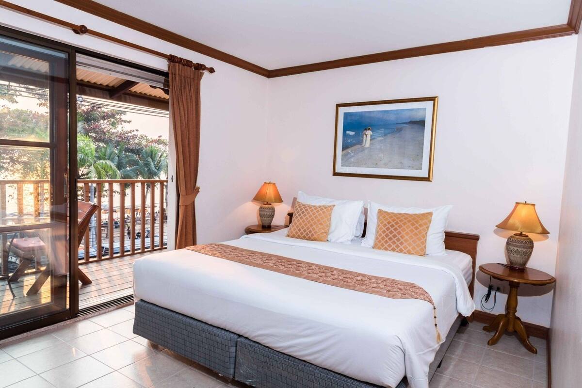 A hotel room with a large bed, two bedside lamps, a painting on the wall, and a balcony with a view of palm trees.