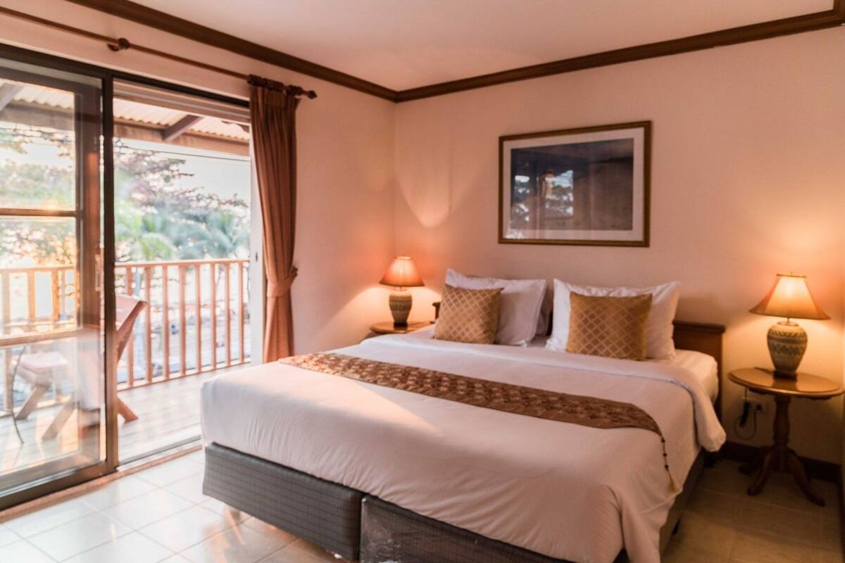 A hotel room with a bed, two bedside lamps, a framed picture on the wall, and a balcony with a chair.