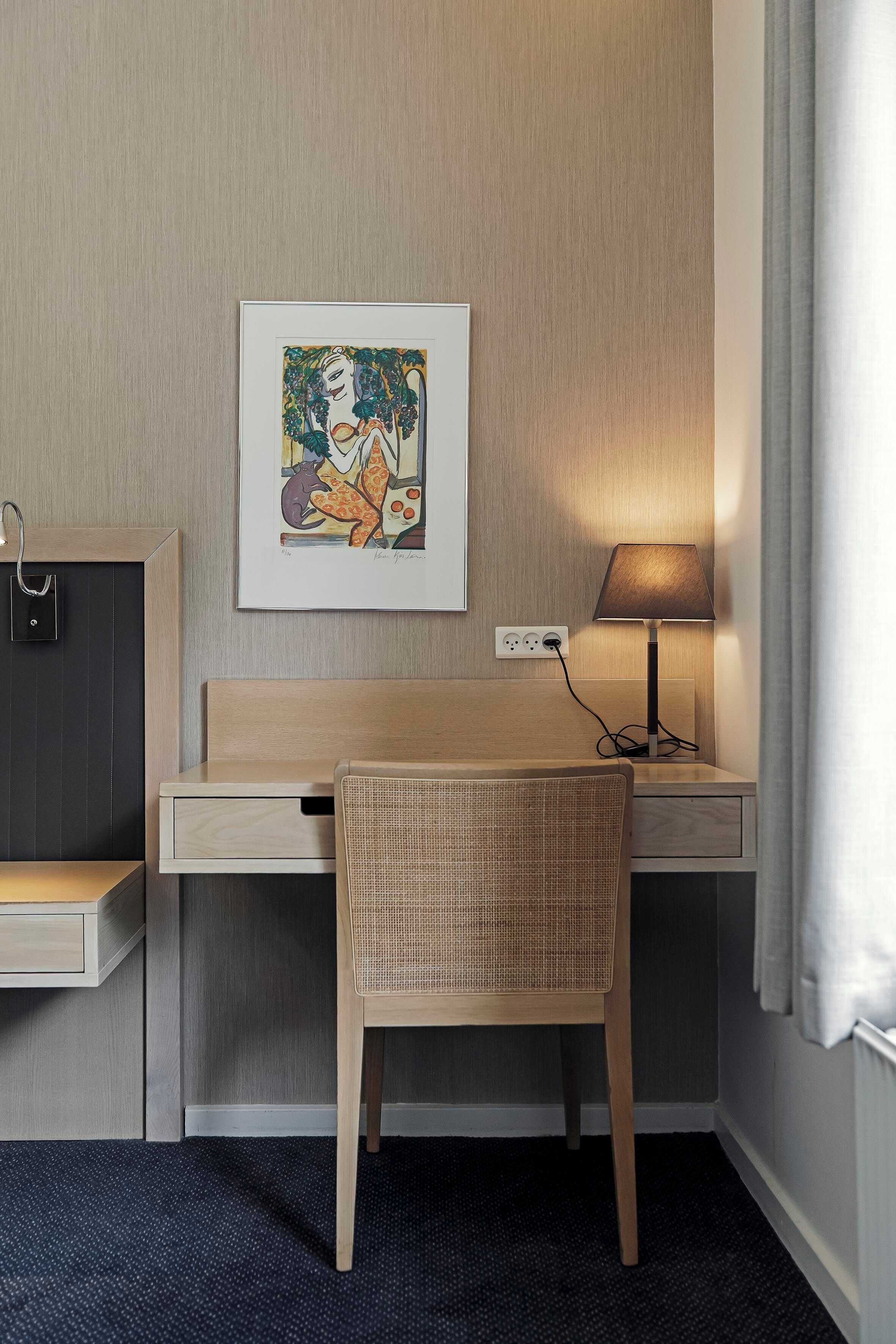 Habitación de hotel con un escritorio de madera, una silla de mimbre, un cuadro en la pared, una lámpara de noche y un enchufe.