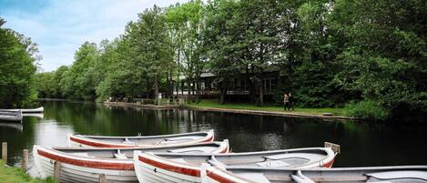 Una fila de pequeñas embarcaciones amarradas a la orilla de un río, con un edificio y árboles al fondo.