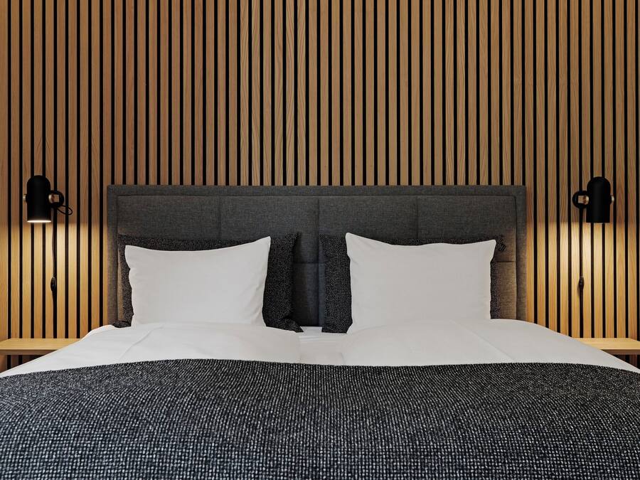 Cama bem arrumada com cabeceira cinza, duas mesas de cabeceira e luminárias fixadas na parede.