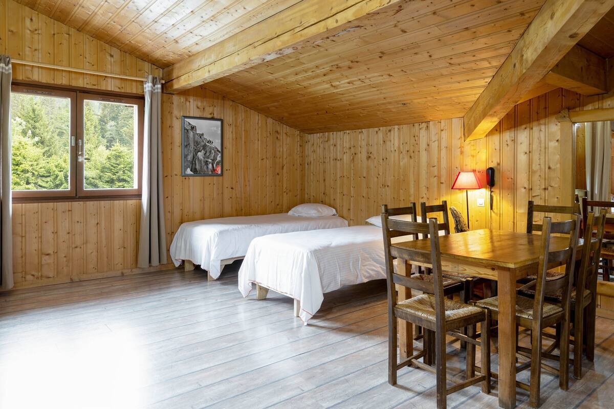 A wooden cabin with two single beds, a dining table with chairs, and a window with curtains.