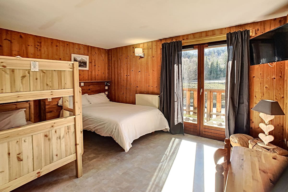 A wooden cabin bedroom with bunk beds, a single bed, a large window with curtains, a wooden dresser, and a TV.
