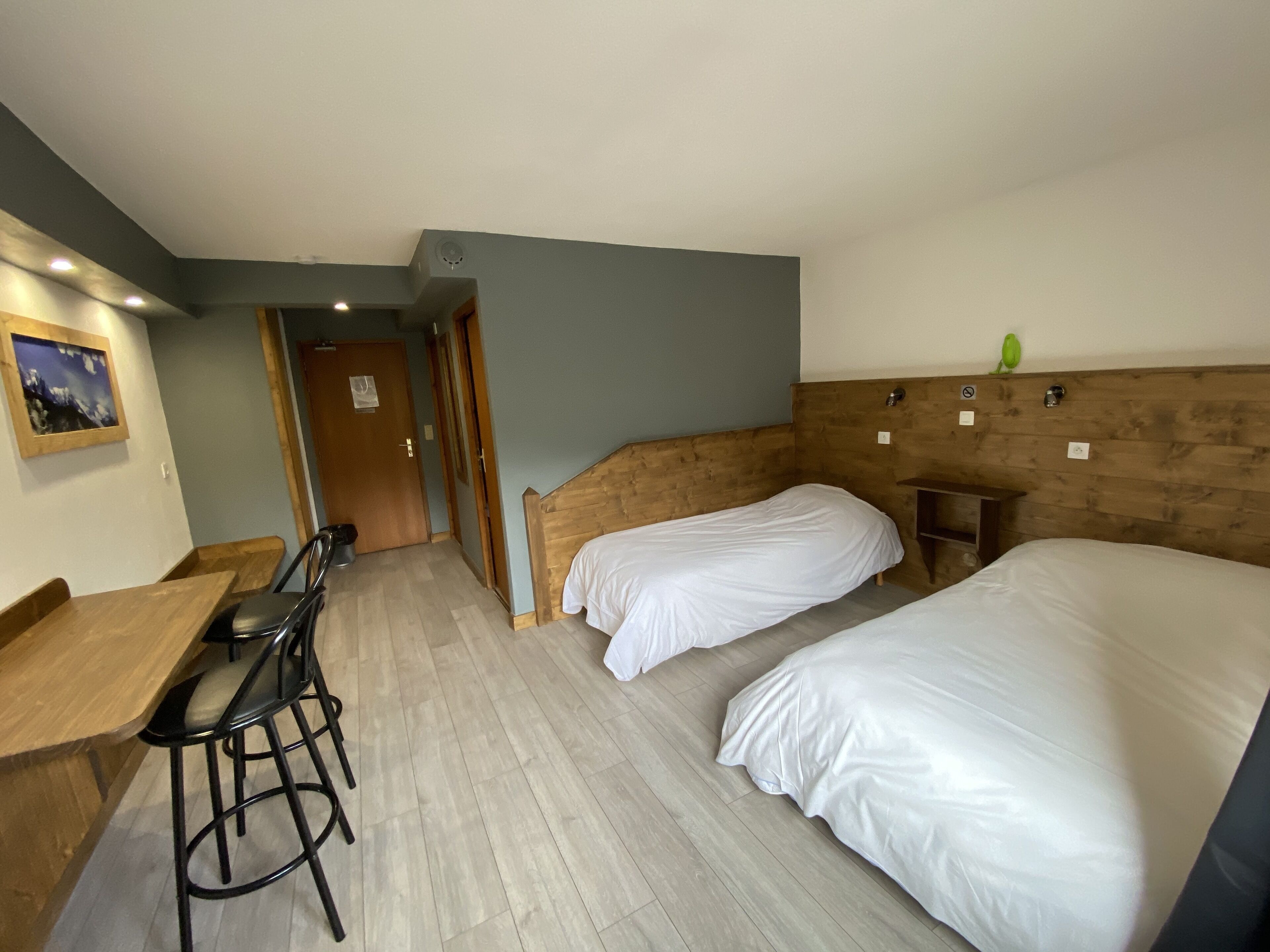 Una habitación con dos camas, un cabecero de madera, un taburete pequeño de madera, un escritorio con una silla y un cuadro en la pared.