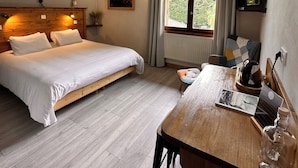 A bedroom with a wooden bed, a wooden desk, a window with curtains, and a view of greenery outside.