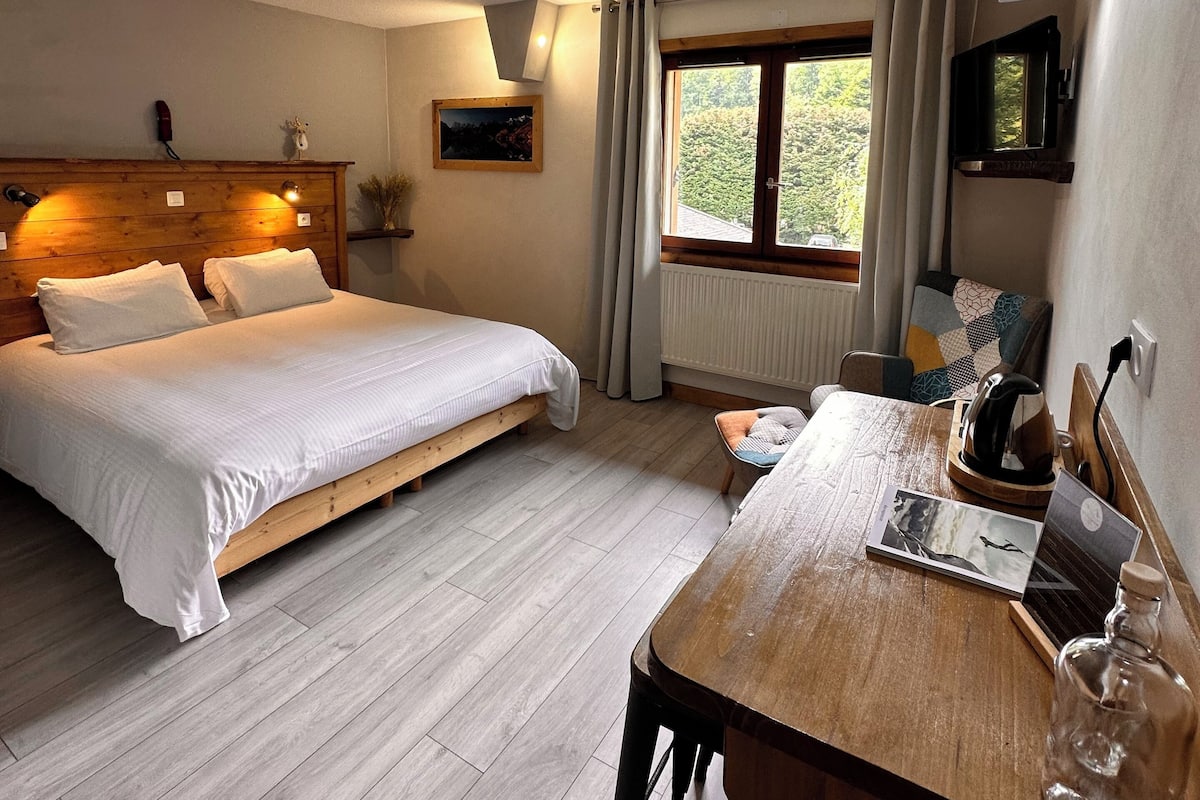 A bedroom with a wooden bed, a wooden desk, a window with curtains, and a view of greenery outside.