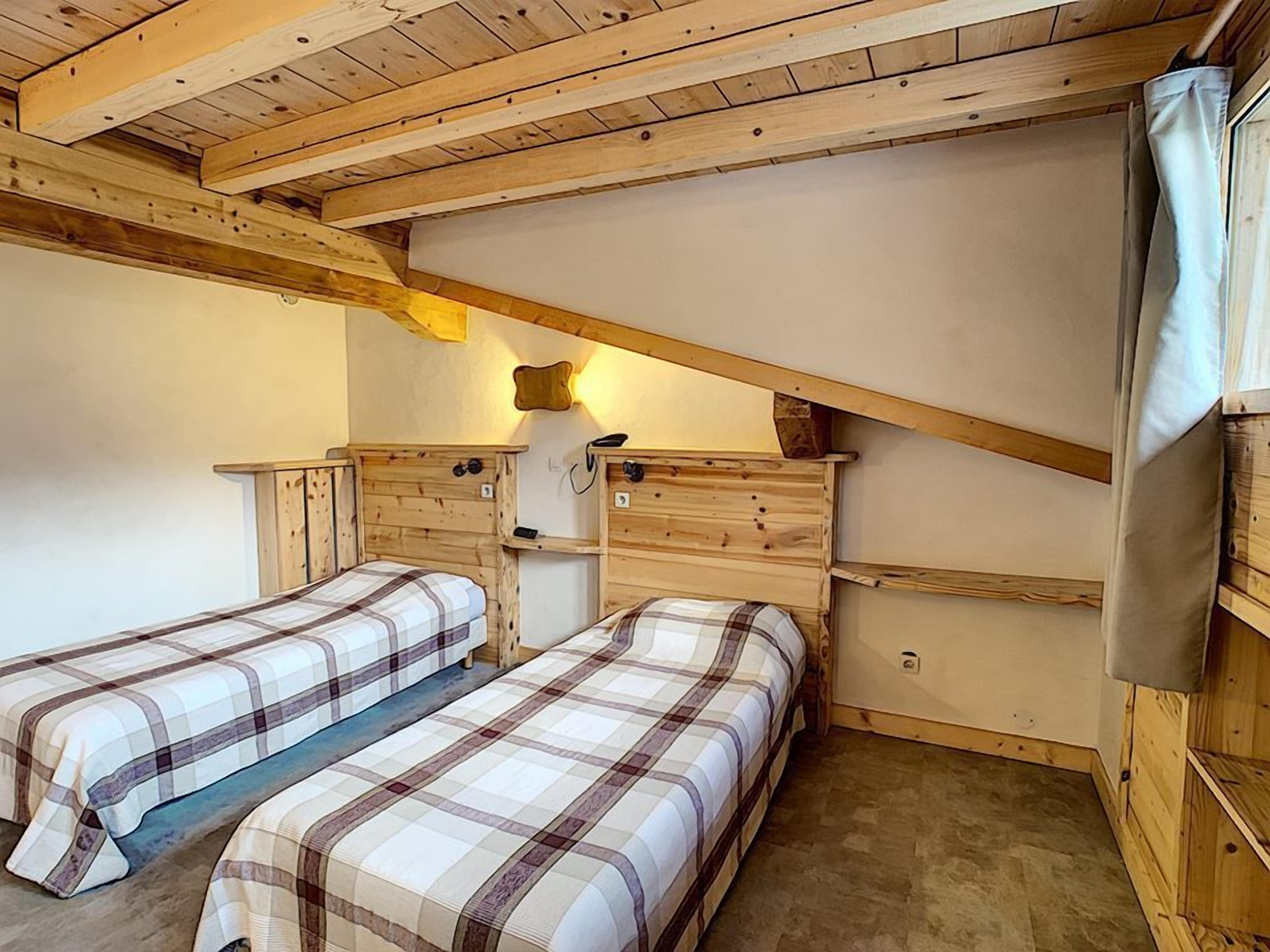 Una habitación acogedora con vigas de madera, dos camas con ropa de cama a cuadros, un escritorio pequeño y una lámpara fijada a la pared.