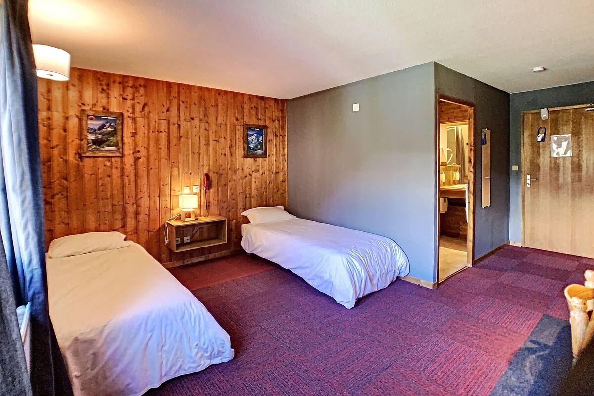 A hotel room with two single beds, a wooden wall, a small bedside table with a lamp, and a door leading to a bathroom.