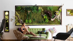 A room with a green sofa, a wicker chair, a black rocking chair, a small round table, and a large moss wall art piece.