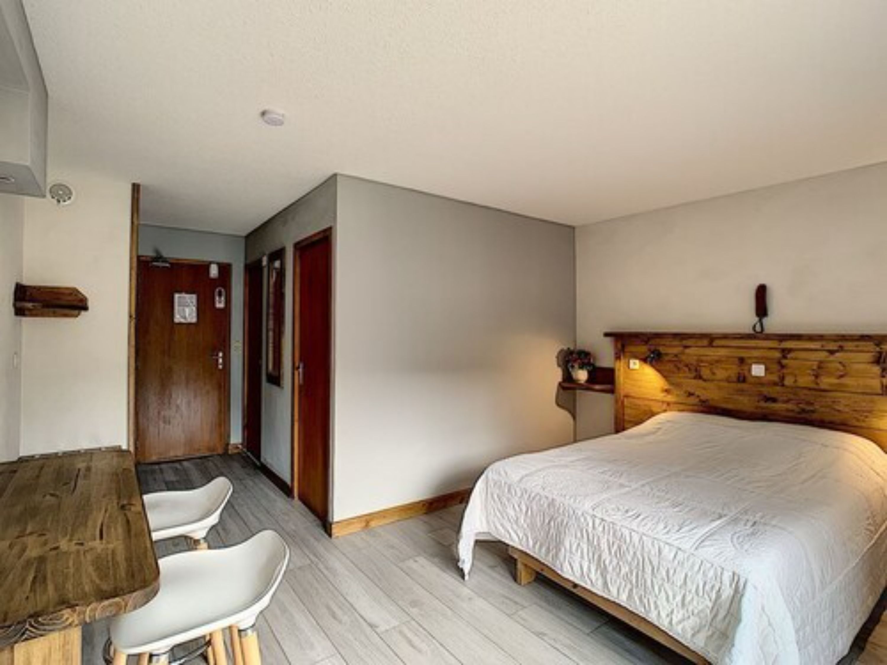 Un dormitorio con cabecera de madera, cama con edredón blanco, un escritorio de madera con dos sillas y una puerta de madera.