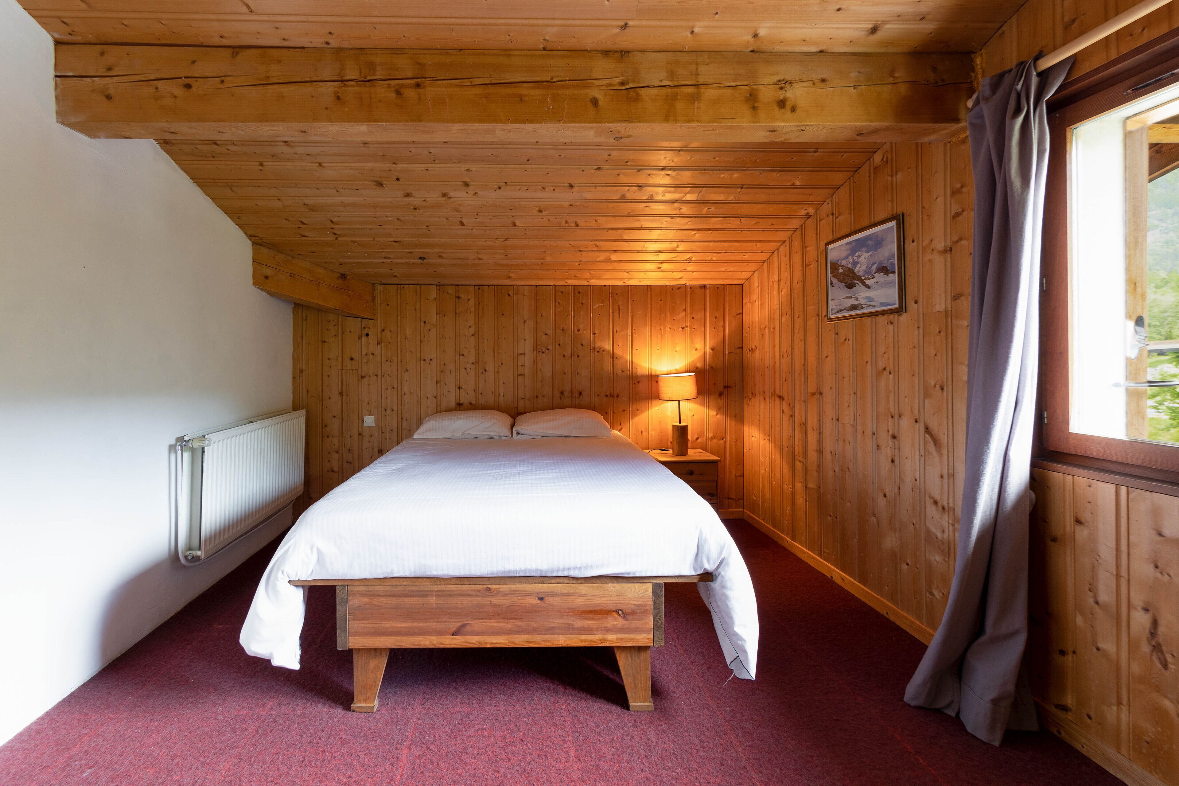 A wooden-paneled room with a bed, a nightstand with a lamp, and a window with curtains.