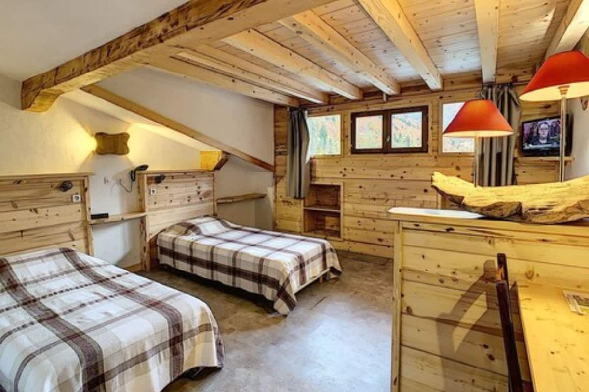 A cozy attic bedroom with two beds, wooden beams, a desk, and a television.