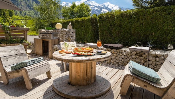 An outdoor dining area with a round wooden table, two wooden benches, and a stone wall.