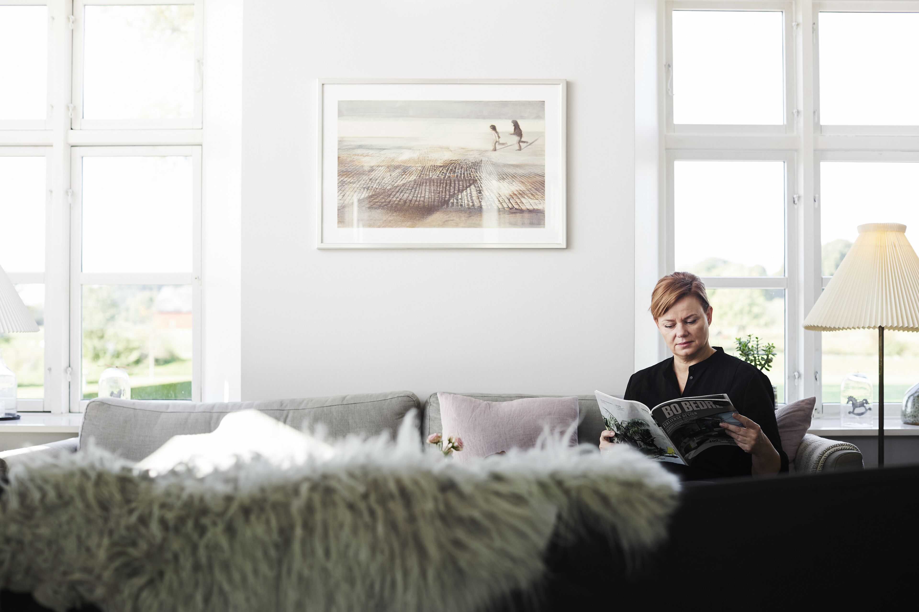 Una persona legge una rivista su un divano grigio con una coperta bianca e soffice, un quadro appeso al muro e una lampada su un tavolino.