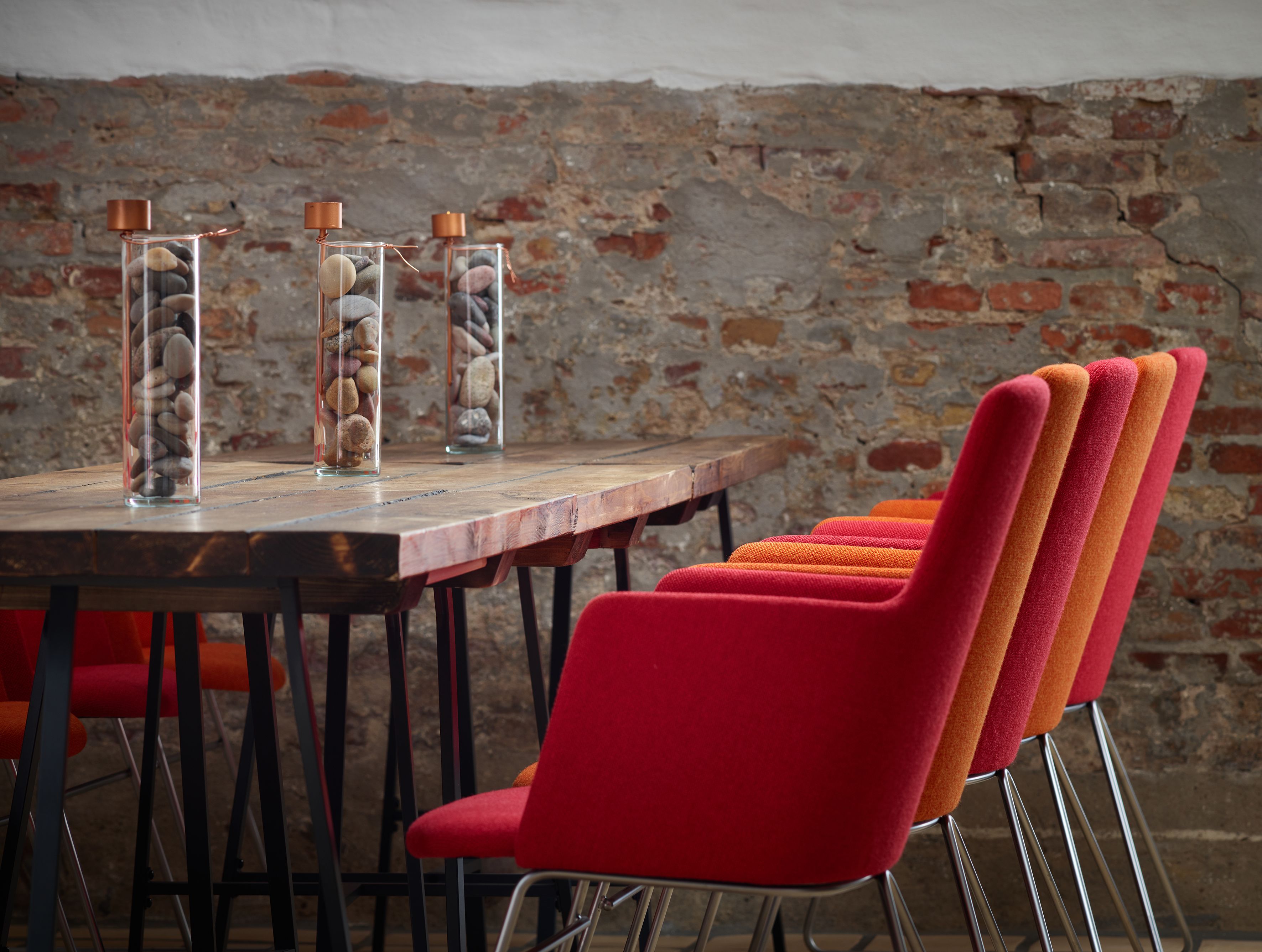 Una mesa de madera con tres jarrones de vidrio que contienen piedras, sillas tapizadas en rojo y un fondo de pared de ladrillo.