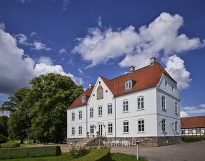 Un edificio grande de color blanco con techo de tejas rojas, múltiples ventanas y base de piedra.