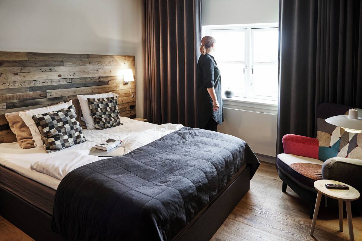 Una habitación de hotel moderna con una cama grande, un cabecero de madera, una ventana con cortinas, una mesita pequeña y un sofá.