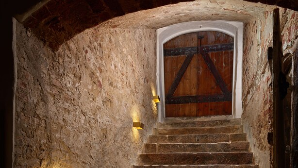 Una escalera de piedra que lleva a una puerta de madera con un portón de metal, iluminada por luces en la pared.