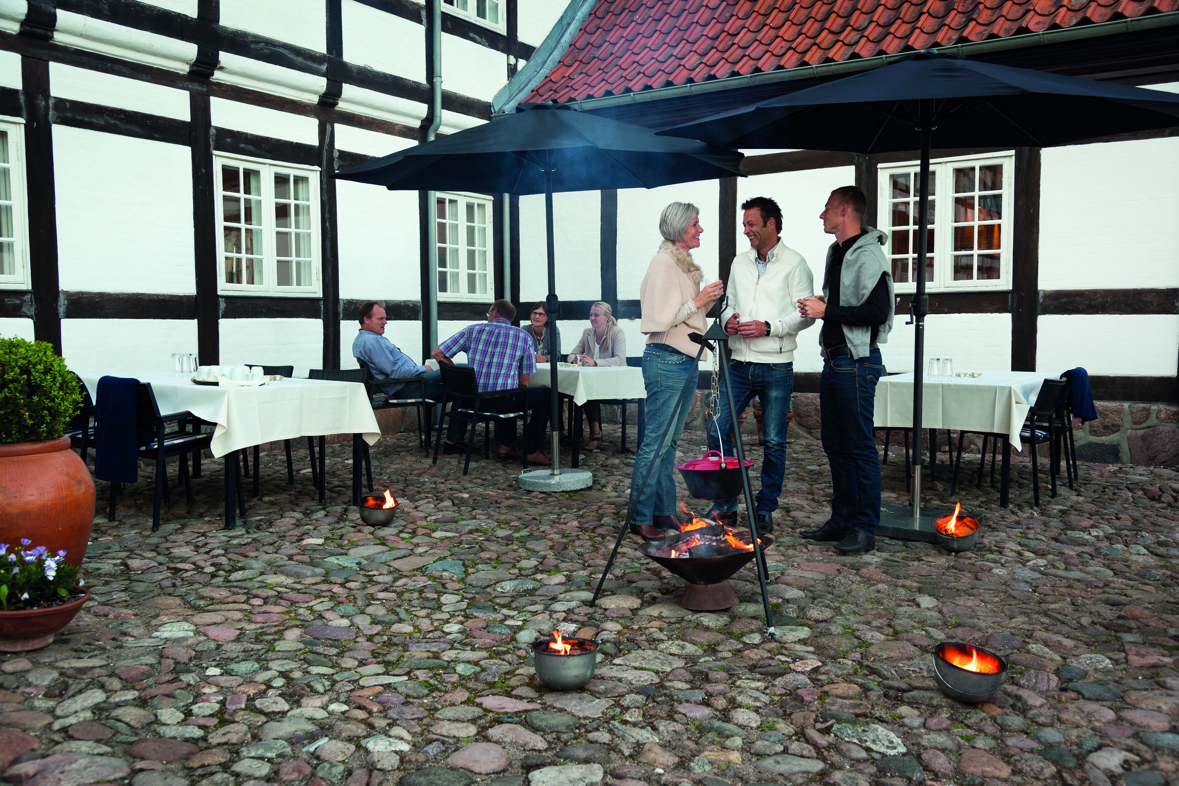 An outdoor dining area with stone flooring, tables with white tablecloths, and people gathered around a fire pit.