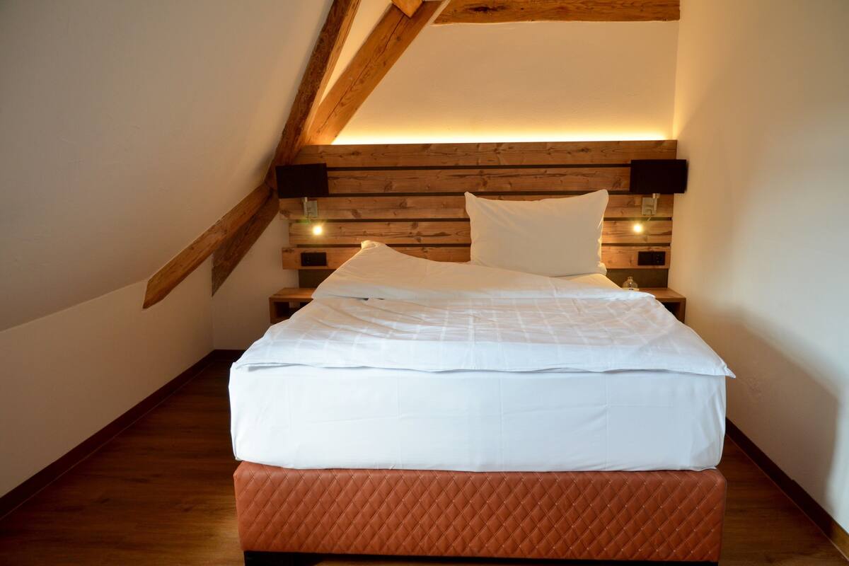 A bedroom with a wooden ceiling, a bed with white bedding, and a nightstand with a lamp.